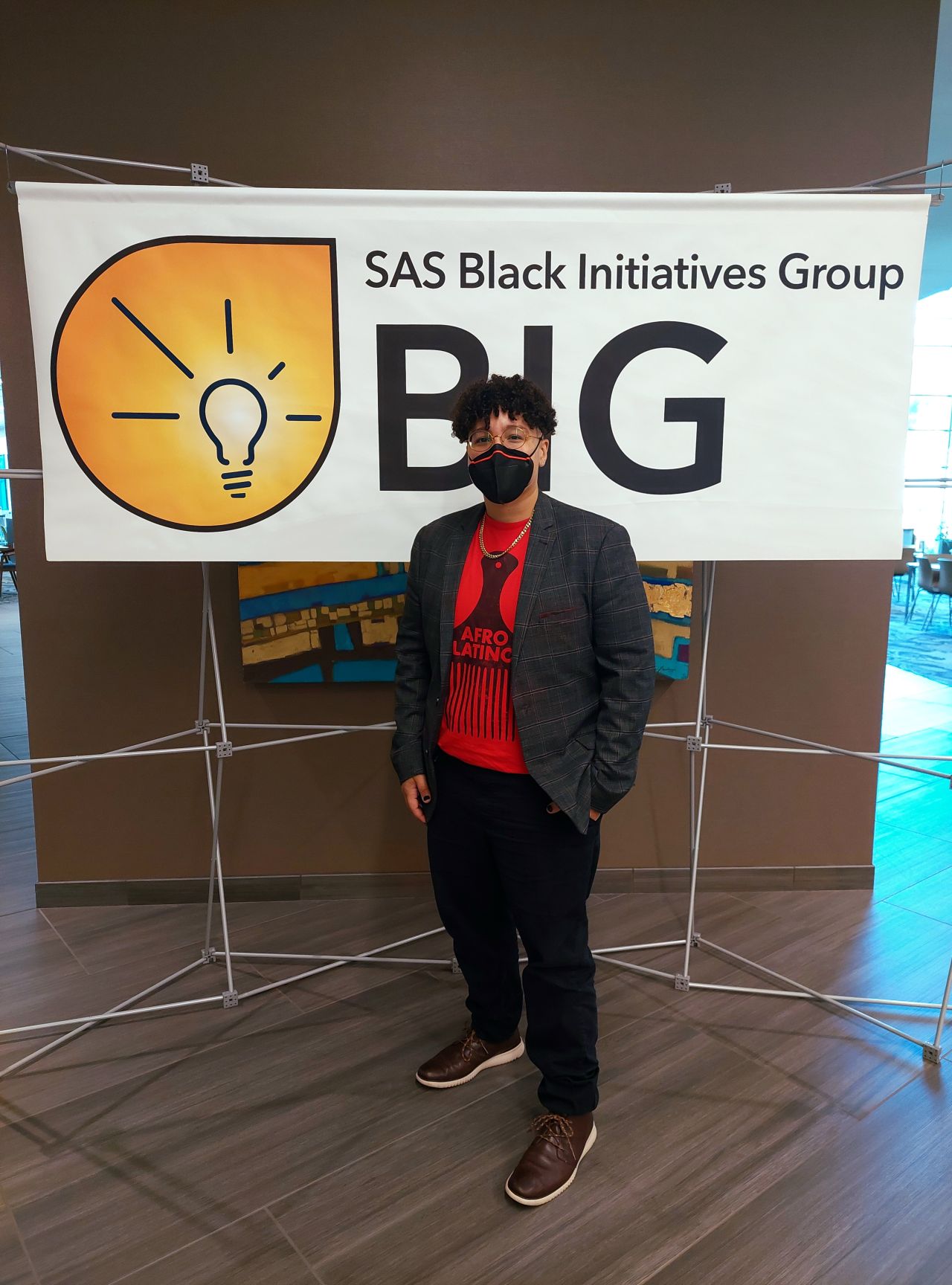 Keslie standing in front of a banner reading 'SAS Black Initiatives Group' while wearing a KN95 respirator and a red shirt that says 'Afro-Latino'.